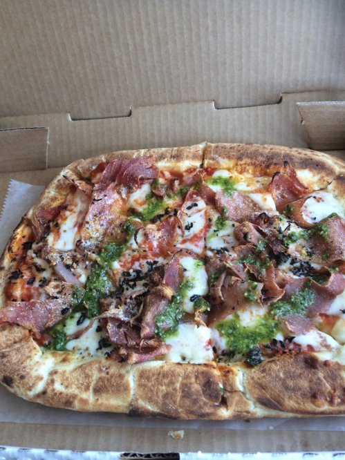 A rectangular pizza topped with cheese, pesto, and slices of cured meat in a cardboard box.